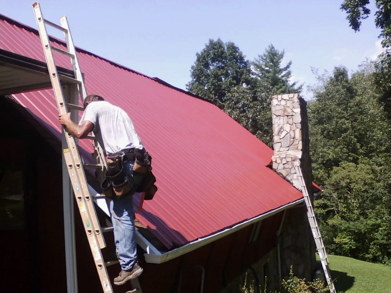 Roofer on a ladder beside a red metal roof and stone chimney — Soffit & Fascia Repair by Roofing Brothers in Arvin